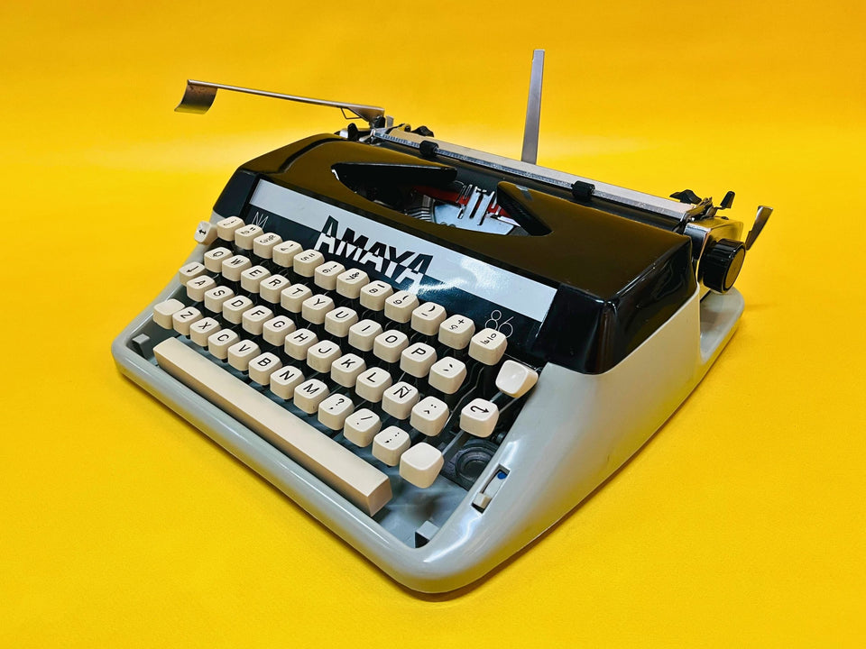 Front-side view of a sleek two-tone Amaya 86 manual typewriter with a glossy black top and grey body against a yellow background