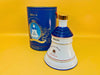 Front view of a white and blue porcelain Bell's Whisky bell-shaped decanter next to its original blue commemorative tube.