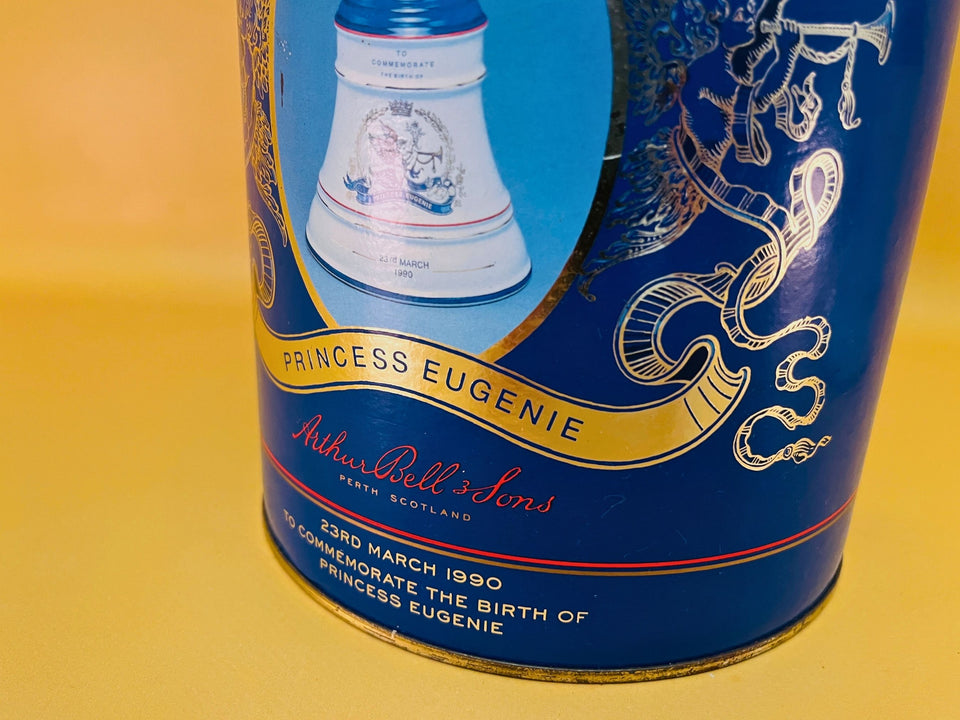 The porcelain decanter, original gold-tone cork stopper, and commemorative cylindrical tin displayed against a yellow background.