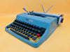 Front view of a classic blue-green Olivetti Lettera 32 manual typewriter in excellent condition with its leather case.