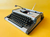 Front-side view of a 1970s AEG Olympia Traveller Deluxe manual typewriter in Arctic White with dark grey side panels against a yellow background.