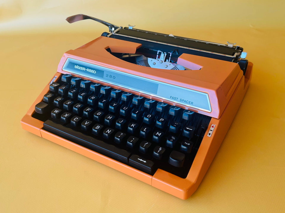 Front view of a striking Safety Orange Silver Reed 280 manual typewriter with its original black transport case against a yellow background.