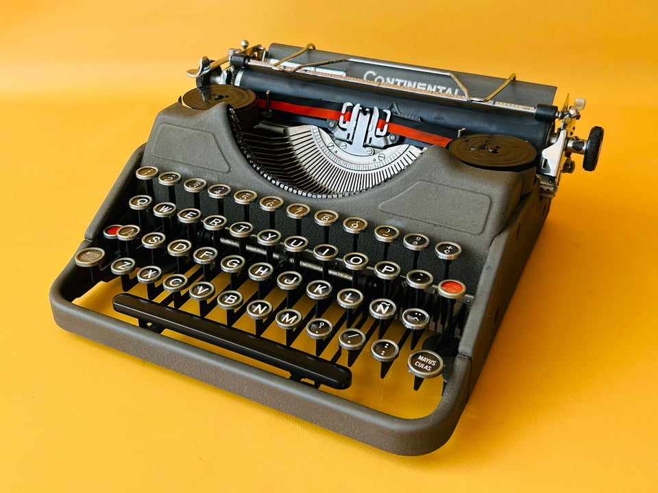 Front-side view of a 1940s matte black Continental Klein Model 100 typewriter with glass keys against a yellow background.