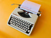 Top-down view of a pristine white Hermes Baby manual typewriter against a vibrant yellow background.