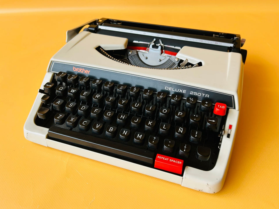 Front-side view of a white Brother Deluxe 250 TR manual typewriter with a black keyboard mask and red repeat spacer against a yellow backgroun