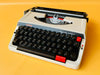 Front-side view of a white Brother Deluxe 250 TR manual typewriter with a black keyboard mask and red repeat spacer against a yellow backgroun