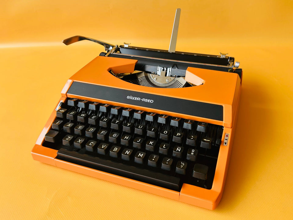Front view of a vibrant Electric Orange Silver Reed manual typewriter with a black keyboard mask against a yellow background