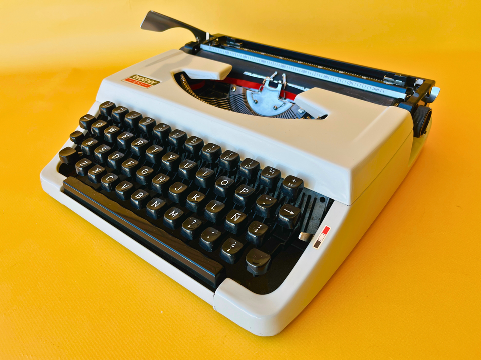 Front view of a crisp Arctic White Brother 210 manual typewriter with its original black hardshell case against a yellow background