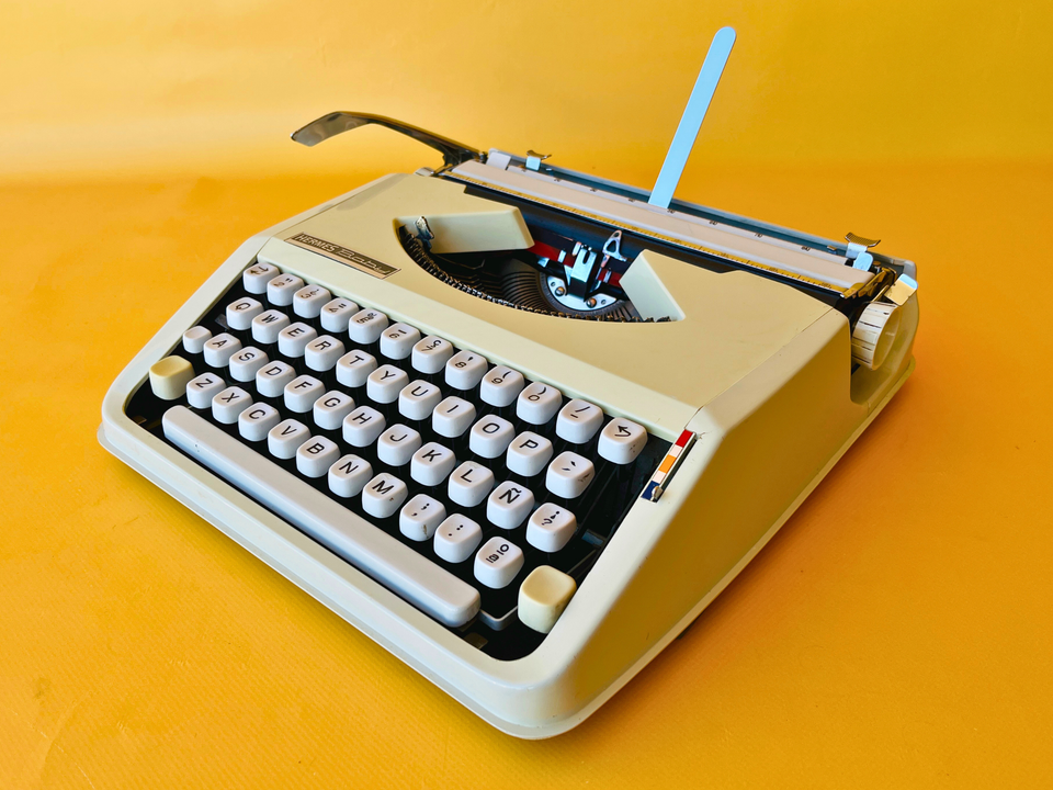 Front view of a 1967 Smooth White Hermes Baby manual typewriter in its ultra-slim metal case against a yellow background