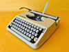 Front view of a 1967 Smooth White Hermes Baby manual typewriter in its ultra-slim metal case against a yellow background