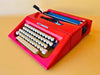 Front-side view of a pristine, vibrant red Olivetti Lettera 25 manual typewriter with crisp white keycaps against a smooth yellow background.