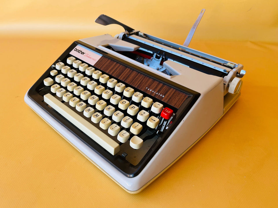Front-angle view of a classic cream and black Brother Deluxe 1300 Tabulator typewriter sitting next to its original grey zippered travel case against a yellow background.