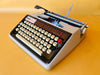 Front-angle view of a classic cream and black Brother Deluxe 1300 Tabulator typewriter sitting next to its original grey zippered travel case against a yellow background.