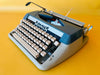 Top-down view of a mint-condition Amaya 86 typewriter featuring a striking two-tone slate blue and light grey body against a vibrant yellow background.