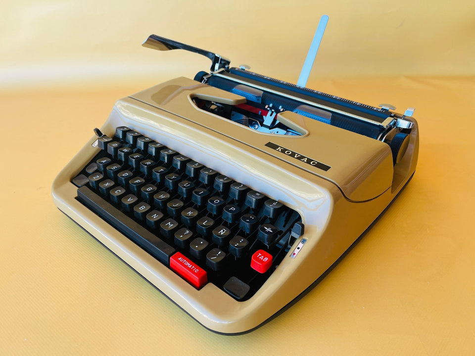Front-side view of a pristine, showroom-condition Kovac Portable typewriter in a high-gloss gold beige finish with black keycaps against a yellow background